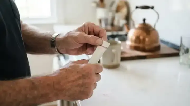 Hands of an older man tearing open a slim single-serve powder stick packet over a kitchen counter, a little pale pink powder visible